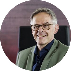 Potrait von Rudi Hoogvliet, der graue Haare hat und auf einem Bürostuhl sitzt. Er trägt einen grauen Anzug und ein schwarzes Hemd sowie eine Brille. Er lächelt und zwinkert.