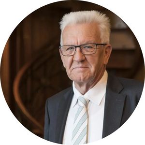 Potraitfoto von Winfried Kretschmann. Er hat weiße Haar, trägt eine rechteckige Brille mit dünnem Rahmen und hat einen grauen Anzug mit weißem Hemd und breit gestreifter Krawatte an.