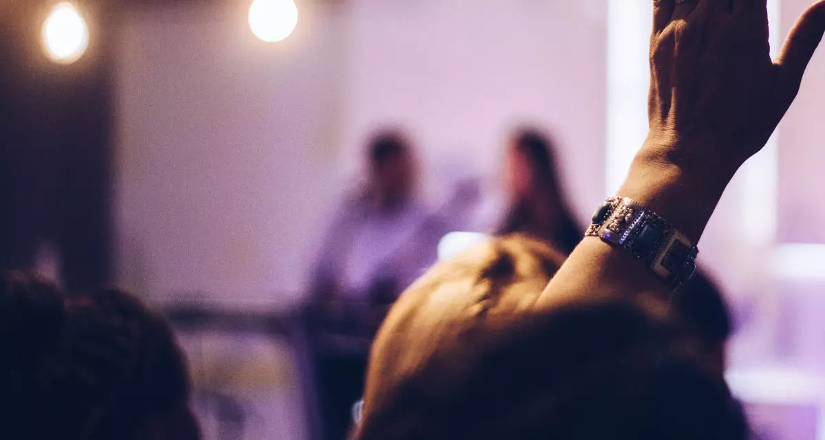 Menschen sitzen auf dem Podium, im Vordergrund eine erhobene Hand.