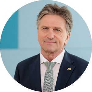 Potrait von Manne Lucha vor blauem Hintergrund. Er trägt ein dunkelblaues Sakko, weißes Hemd mit Krawatte und lächelt leicht.