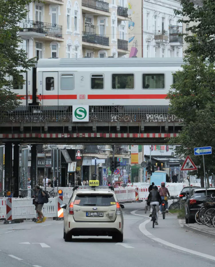 Ein Taxi fährt unter einer S-Bahn-Brücke durch, über die ein ICE fährt. Neben dem Taxi Fahrräder und Fußgänger. Foto: Unsplash|Riiana Izzietovo