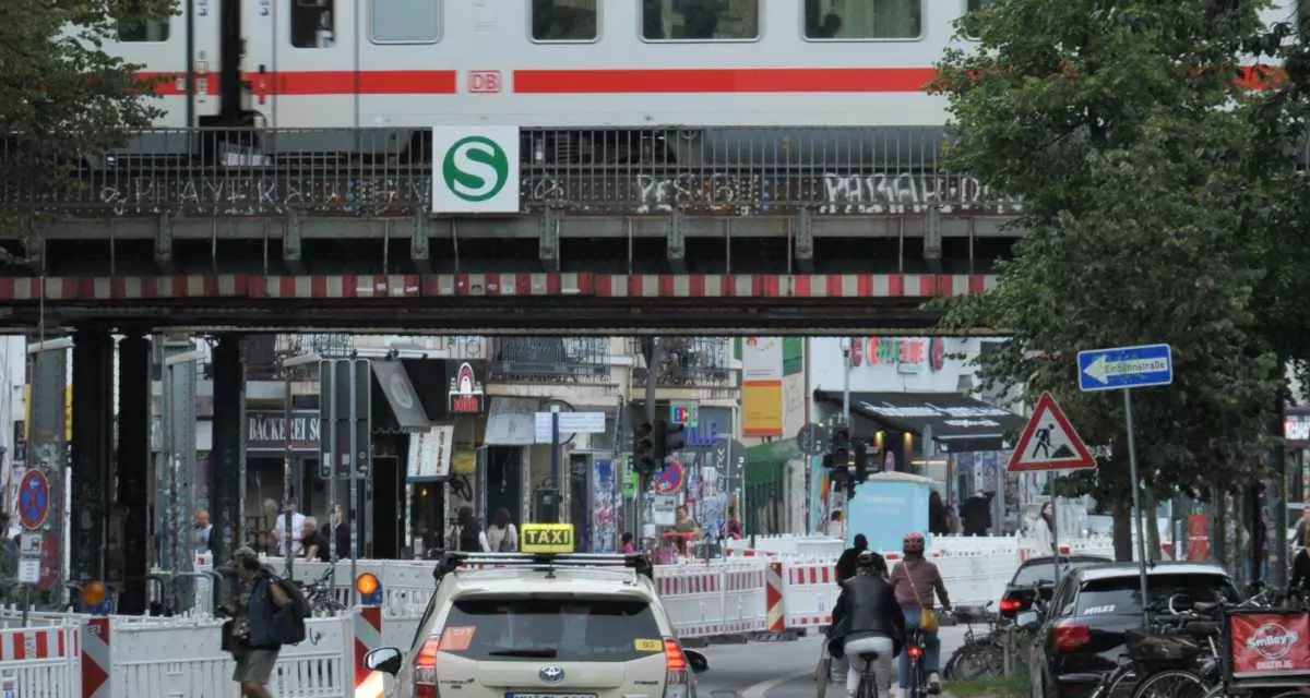 Taxi, Radfahrer und Fußgänger unter einer S-Bahn-Brücke, über die ein ICE fährt.