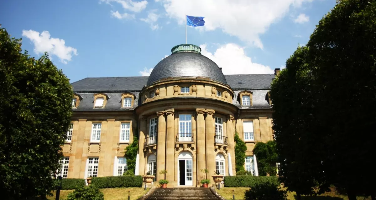 Villa Reitzenstein. flickr | Landesregierung Baden-Württemberg Villa Reitzenstein in Frontalansicht und Sonnenbeschienen. Sandsteinfarbenes Gebäude mit grauem Dach. Vorne in der Mitte runde Kuppel mit EU-Flagge.
