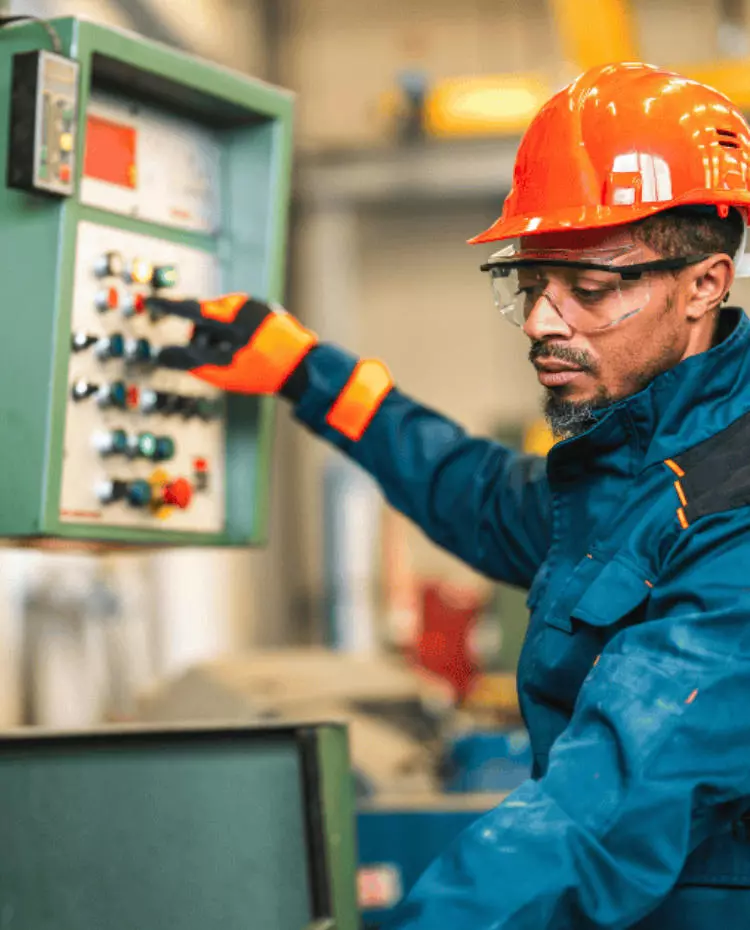 Eine Person im Blaumann mit roten Schutzhelm an einer Maschine. Foto: Canva